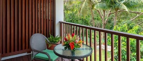 A balcony with a wicker chair, a small round table with a flower vase, and a view of tropical greenery.