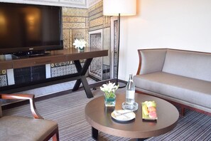 A hotel room with a flat-screen TV, a round coffee table with a water bottle, fruit, and a small vase of flowers. - Hotel Tui Blue Oceana Suites (Hammamet)