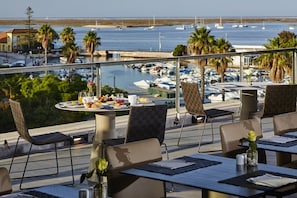 A terrace with tables and chairs, overlooking a marina with boats and palm trees. - Hotel Faro and Beach Club (Faro)