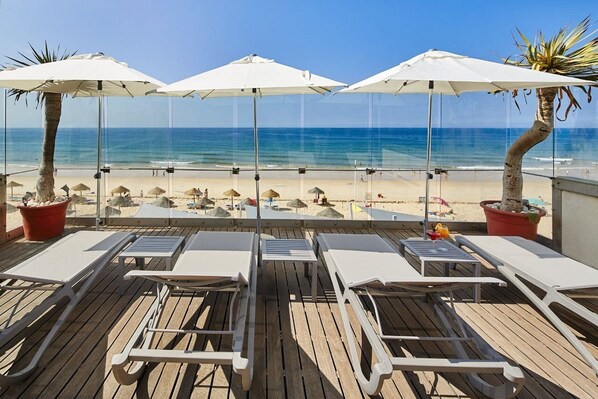 Une terrasse au bord de la plage, dotée de chaises longues, de parasols et offrant une vue sur l’océan.