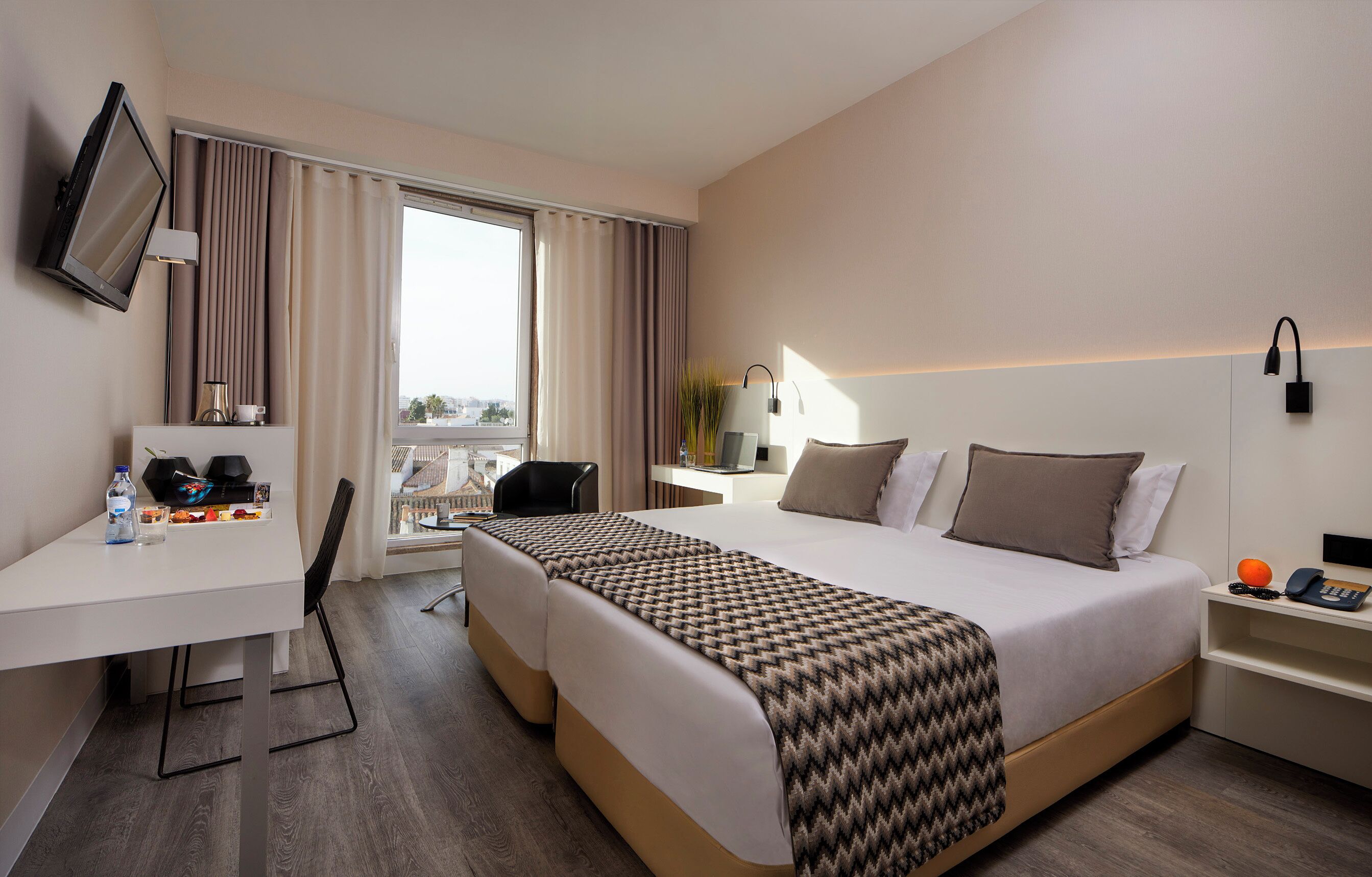 A hotel room with a bed, a desk, a chair, a television, a window with curtains, and a bedside table with a lamp and a phone.
