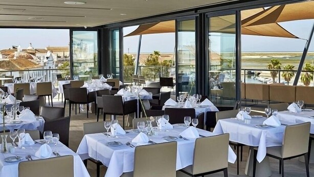 A restaurant with tables set for dining, large windows, and a view of the landscape. - Hotel Faro and Beach Club (Faro)