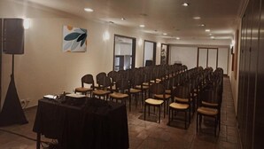 Uma sala de conferências com fileiras de cadeiras, uma mesa com equipamento de som e um orador à esquerda.