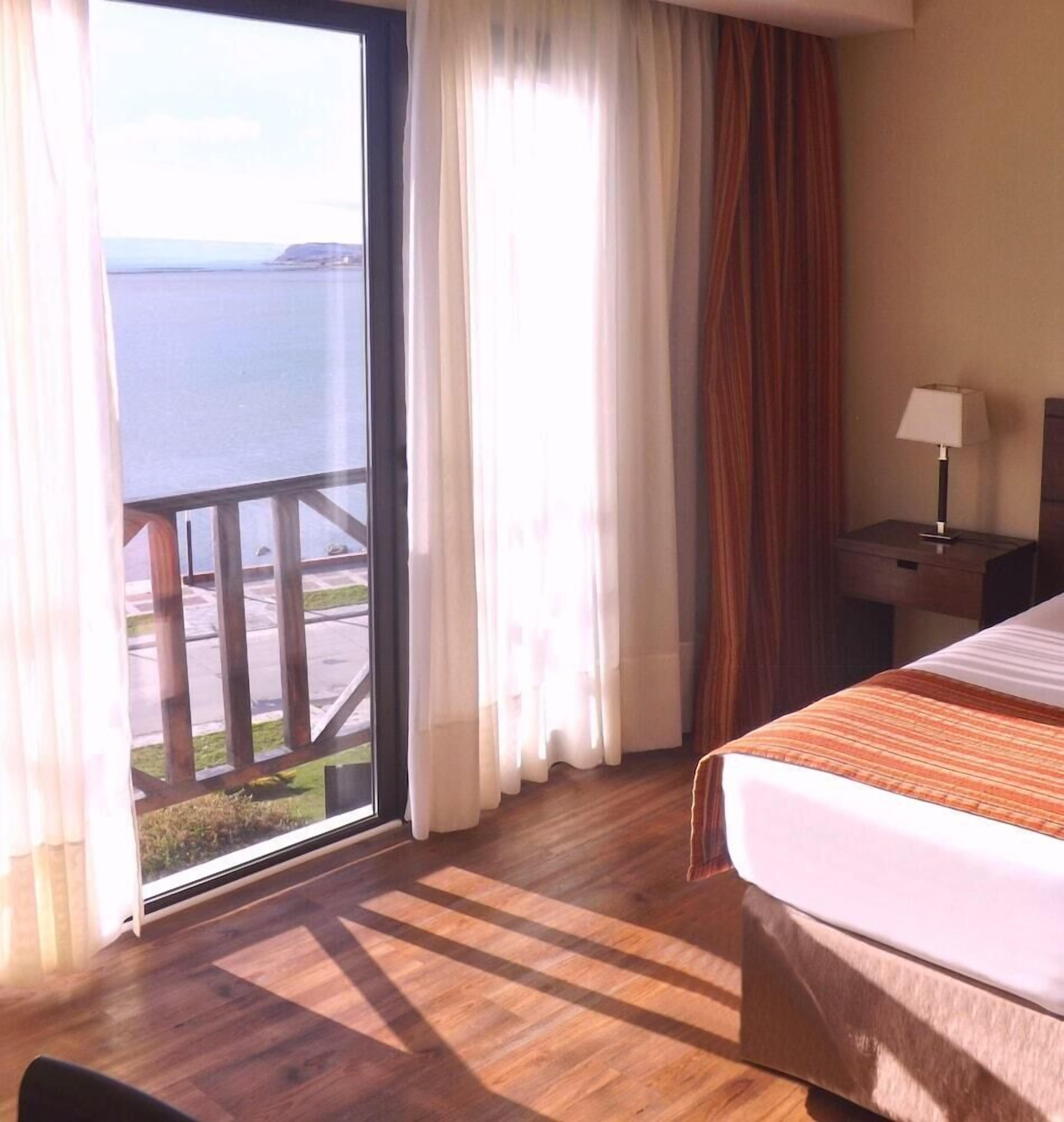Une chambre d’hôtel avec un lit, une table de chevet, une lampe, une fenêtre avec des rideaux et une vue sur la mer.