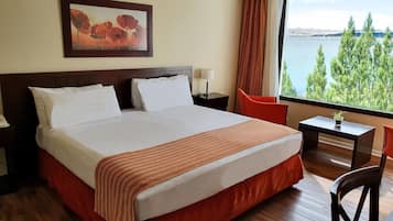 A hotel room with a large bed, a desk, a chair, a nightstand, a lamp, a window with a view of trees and water, and a painting on the wall.