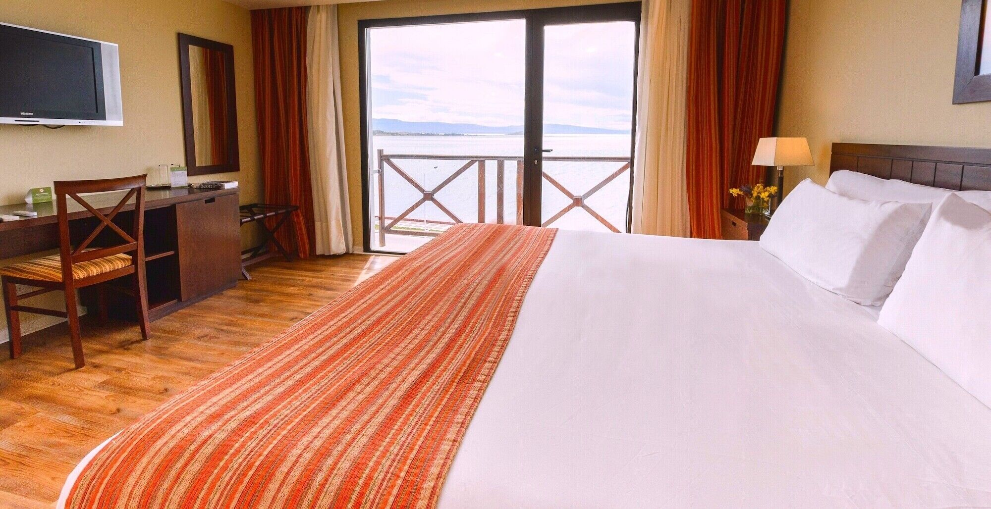 A hotel room with a bed, a desk, a chair, a TV, and a view of the sea.