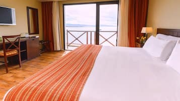 A hotel room with a bed, a desk, a chair, a TV, and a view of the sea.