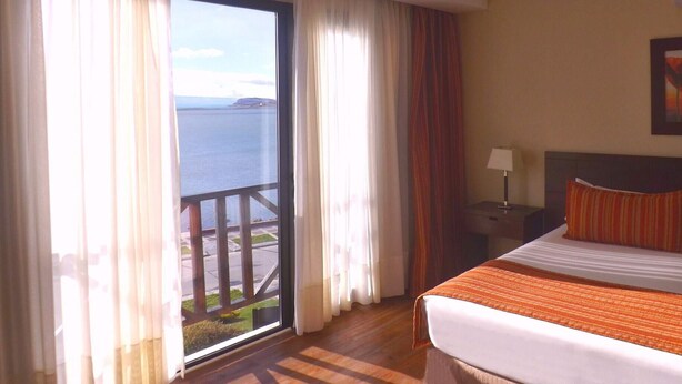 Quarto de hotel com uma cama, mesa de cabeceira, abajur e vista para o mar através de uma janela grande.