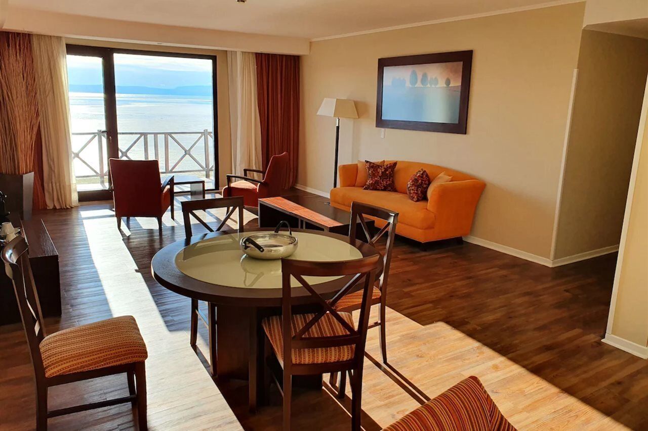 A hotel room with a dining area, a sofa, a TV, and a view of the sea.