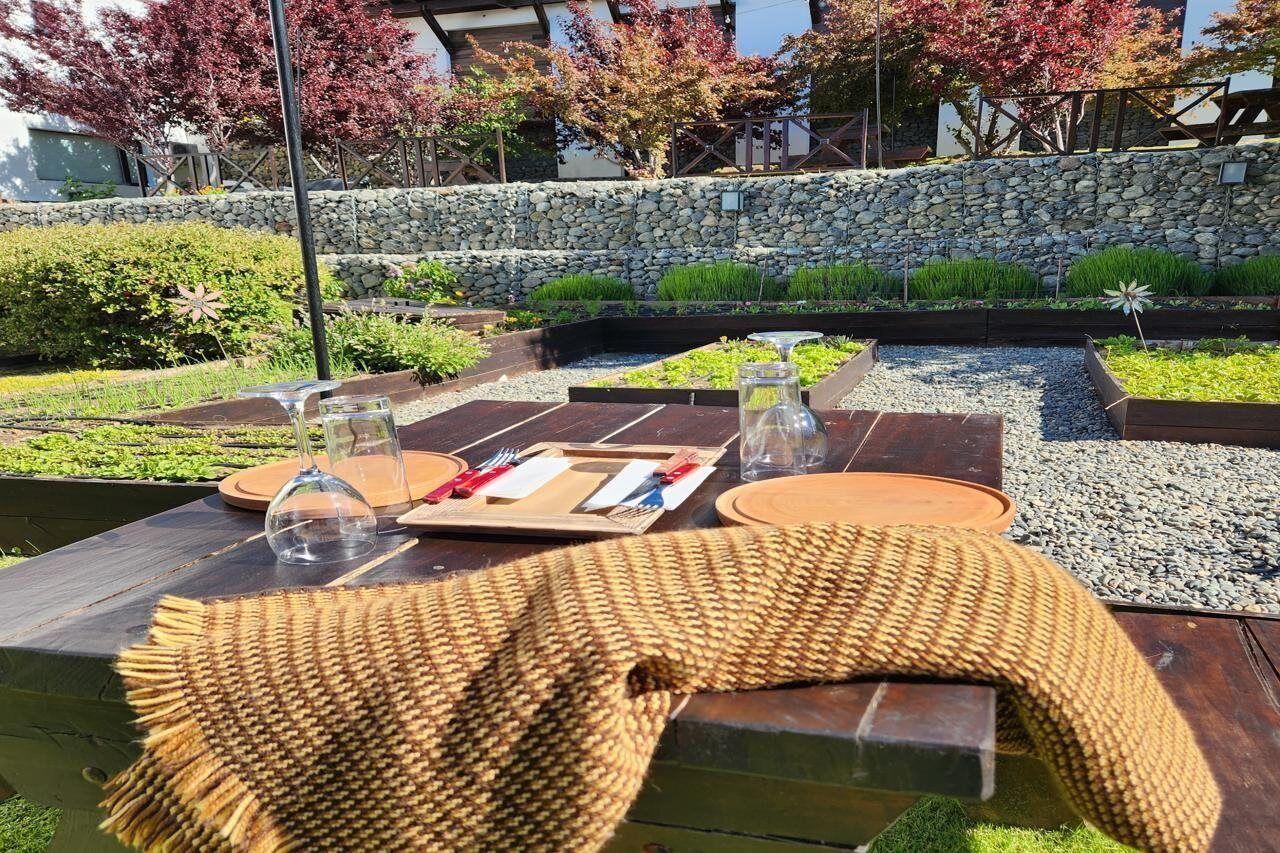 Ein Essbereich im Freien mit einer Steinwand, viel Grün und einem Holztisch für zwei Personen, gedeckt mit Tellern, Besteck und Gläsern.