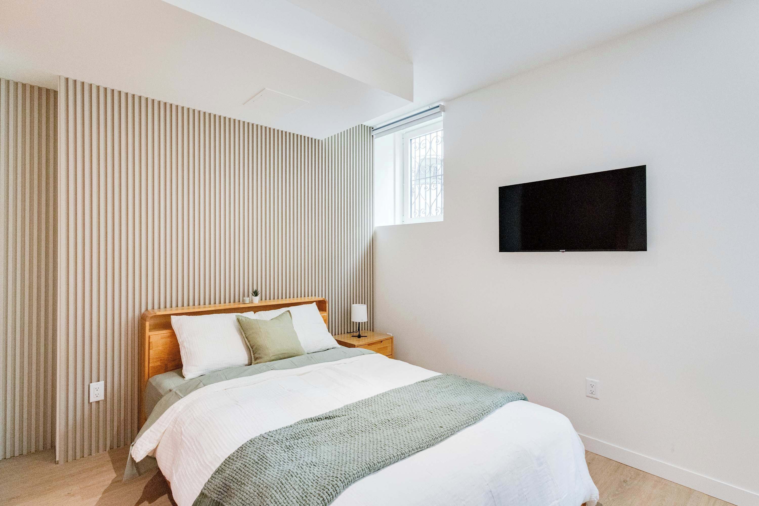 A bedroom with a bed, a nightstand, a TV mounted on the wall, and a window with a view.