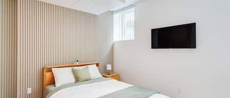 A bedroom with a bed, a nightstand, a TV mounted on the wall, and a window with a view.