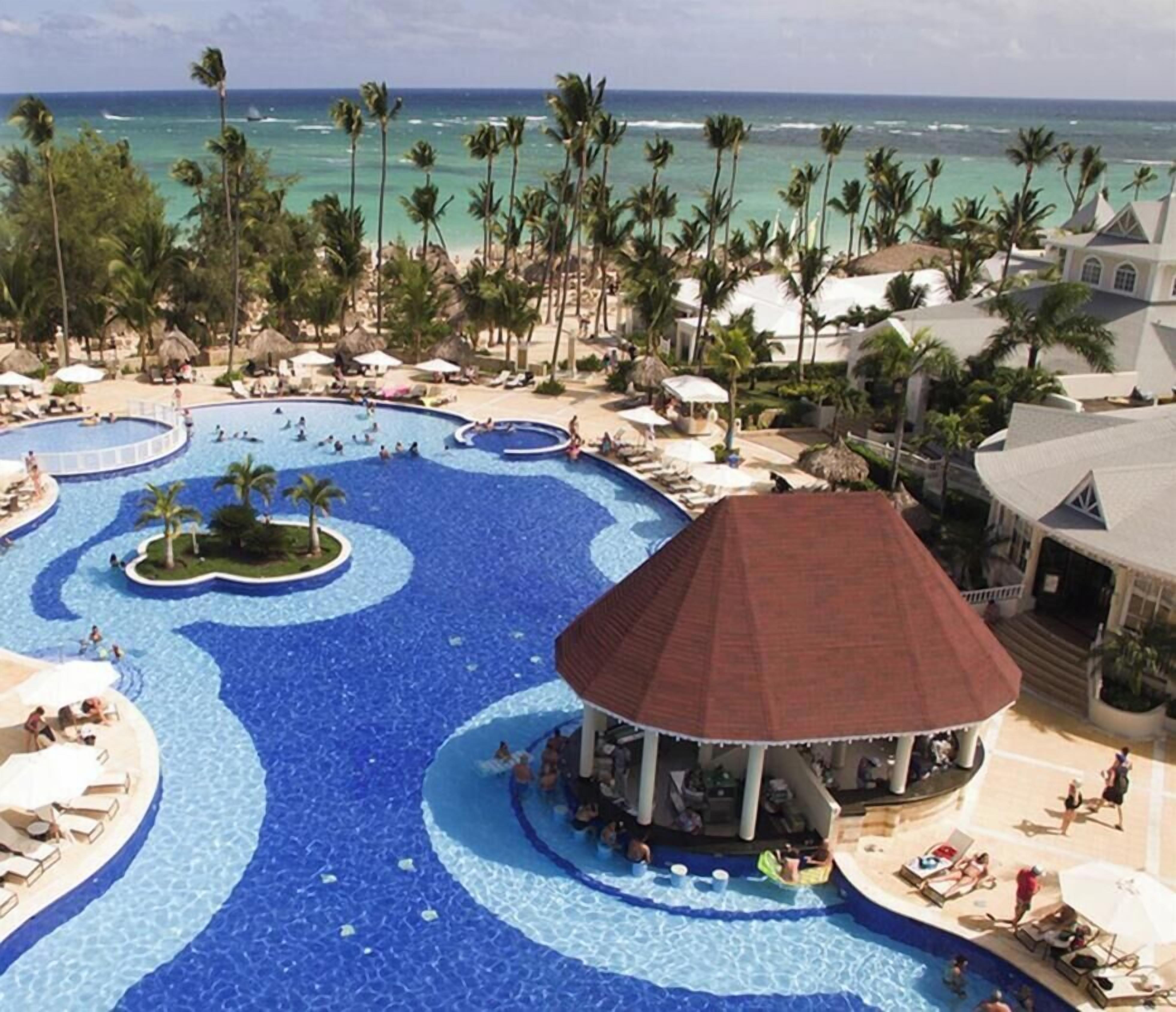 A resort with a large central pool, palm trees, and a beach view.