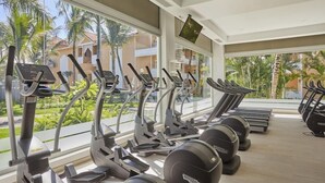 Uma academia com esteiras e equipamentos elípticos, grandes janelas com vista para palmeiras e prédios.
