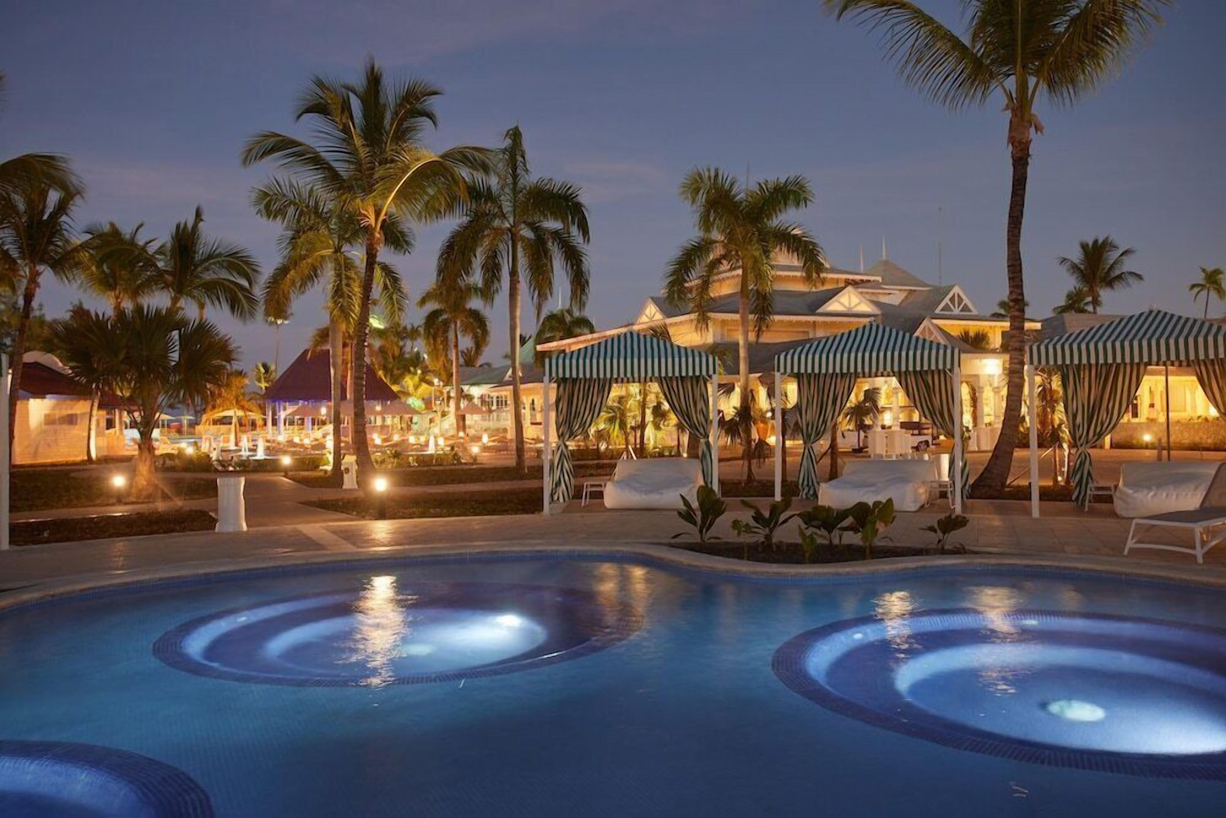 A pool area with lounge chairs and umbrellas, surrounded by palm trees and a building with a distinctive roof.