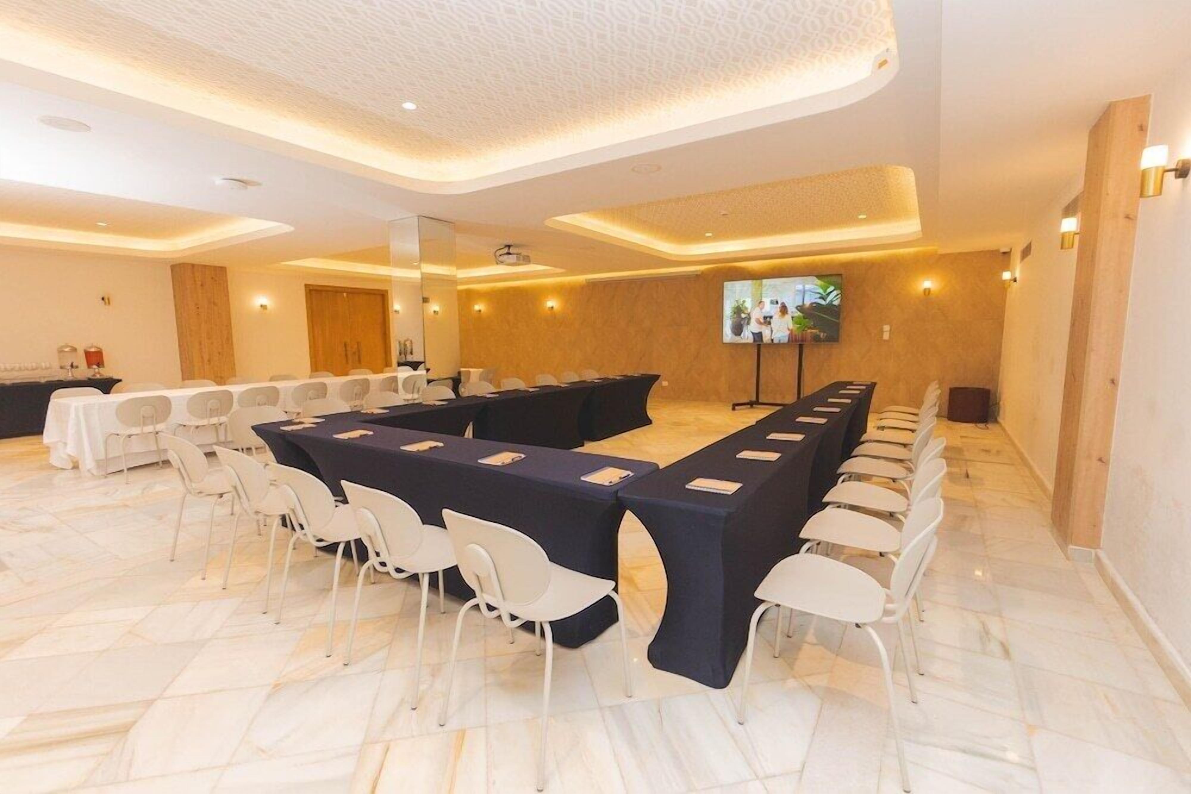 Una sala conferenze con un lungo tavolo, sedie bianche e uno schermo televisivo.