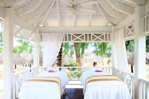 A covered outdoor seating area with two beds, a ceiling fan, and a view of palm trees and beach umbrellas. - Bahia Principe Explore Esmeralda (Punta Cana)