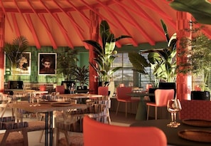Een eethoek binnen met meerdere tafels en stoelen, grote potplanten en lijstwerken aan de wanden.