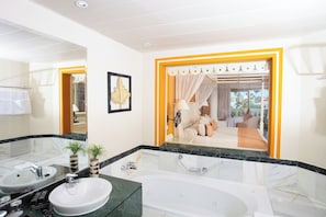 A bathroom with a large mirror, two sinks, a bathtub, and a view into a bedroom with a bed and curtains. - Bahia Principe Explore Esmeralda (Punta Cana)