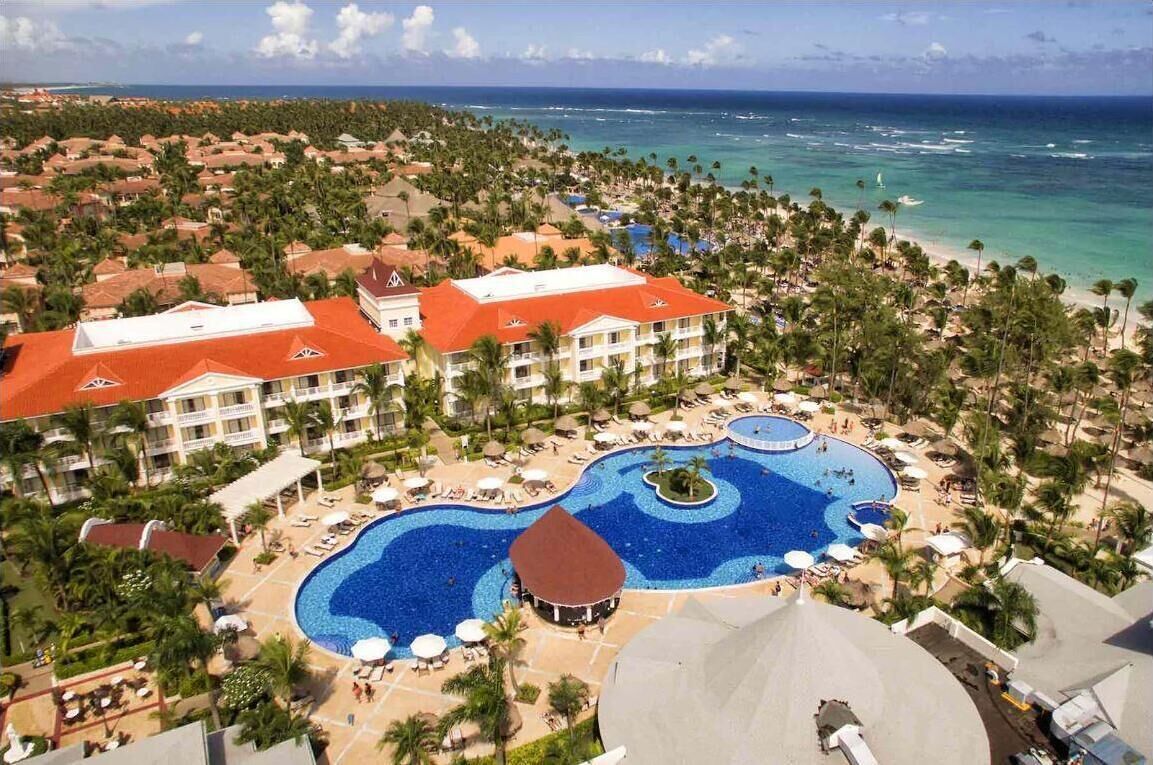 A resort with a large central pool, multiple buildings with red roofs, and a beach area.