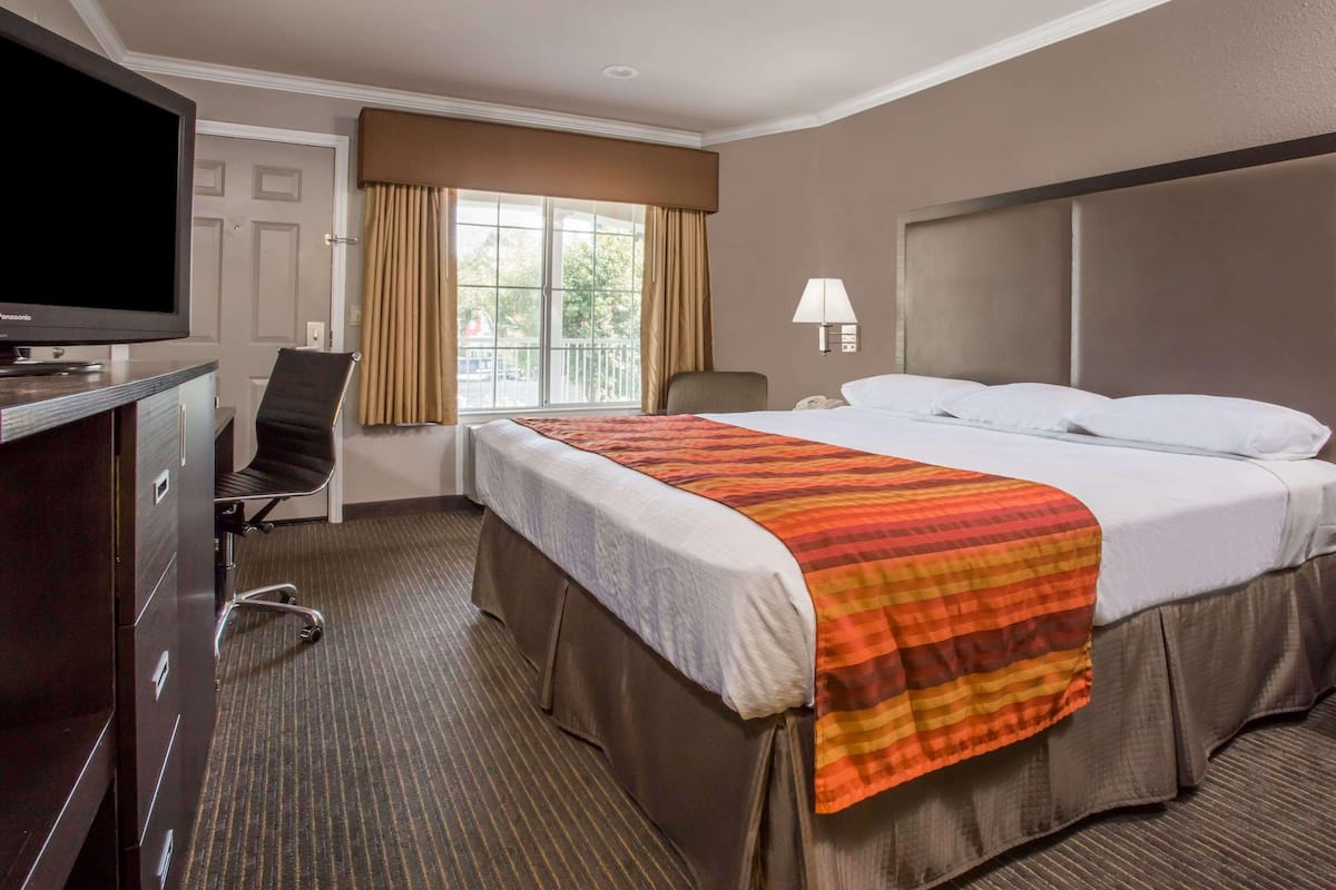 Habitación de hotel con cama, escritorio con televisor, silla, ventana con cortinas y lámpara.