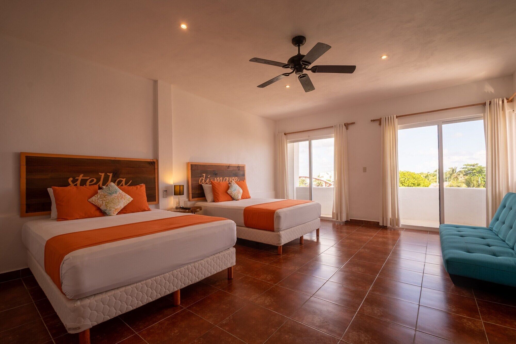 Habitación de hotel con dos camas, ventilador de techo, balcón con cortinas y vista a zonas verdes.
