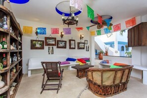 A room with a wooden bookshelf, a white sofa, a wooden chair, a small table, and colorful paper decorations. - Matan Kaan by Guruhotel (Mahahual)