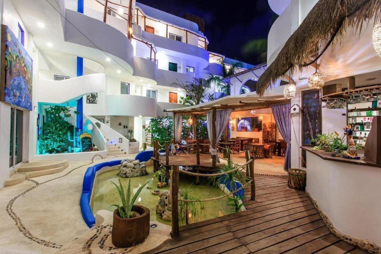 Interior de un hotel de varios niveles con zona de piscina, plantas de interior y un bar.