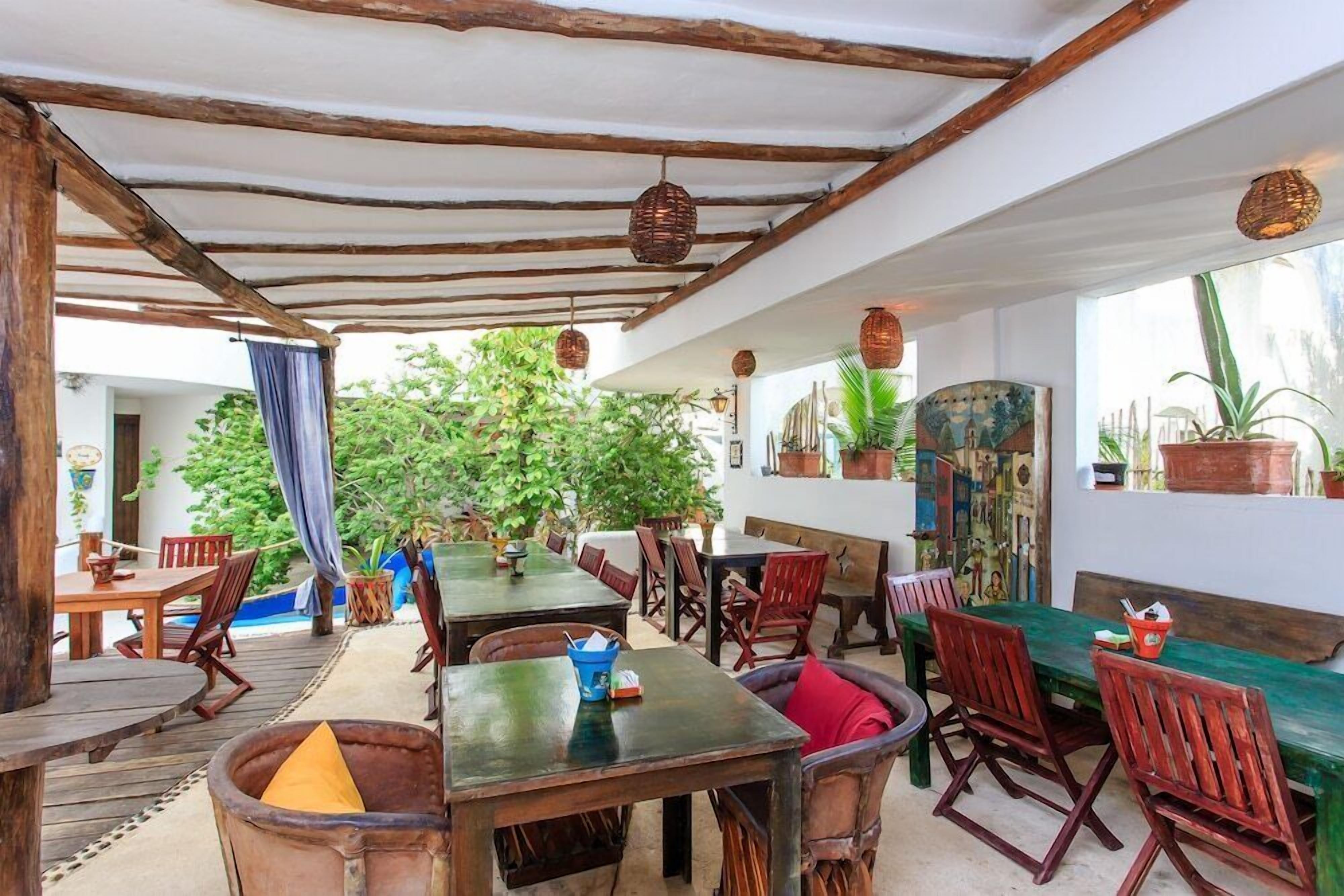 An outdoor dining area with wooden tables and chairs, surrounded by potted plants and artwork.