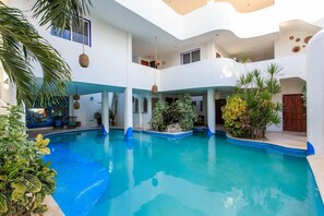 Un área de piscina con piscina azul cristalina, rodeada de paredes blancas y detalles azules, plantas y adornos colgantes.
