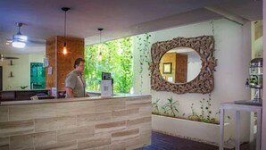 A reception area with a wooden reception desk, a large mirror with an ornate frame, and a view of greenery through the window. - The Palm at Playa (Playa del Carmen)