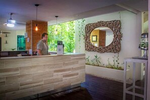 A reception area with a wooden reception desk, a large mirror with an ornate frame, and a view of greenery through the window. - The Palm at Playa (Playa del Carmen)