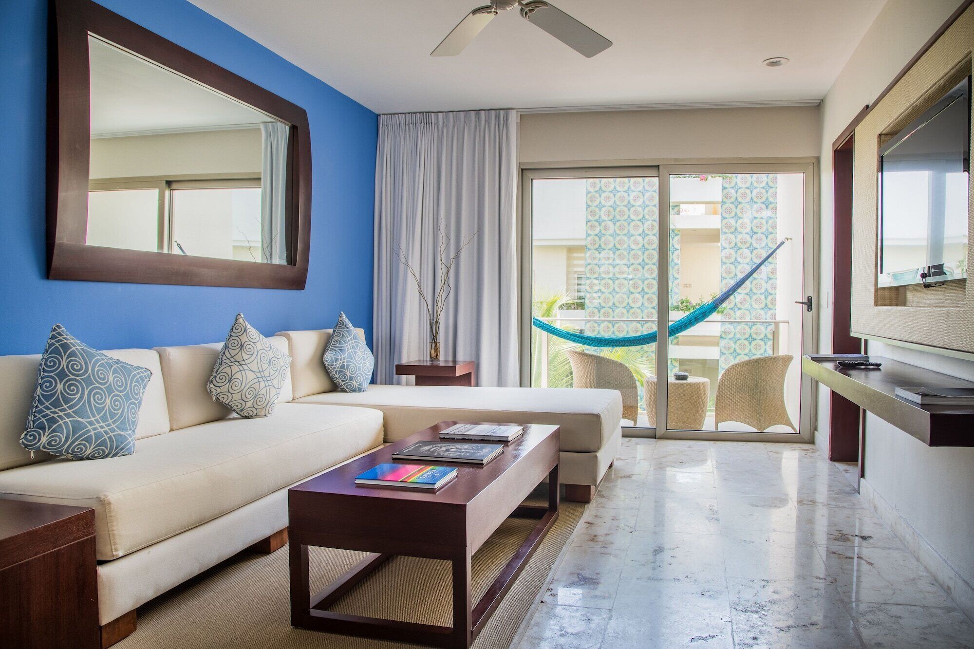 A hotel room with a sofa, a coffee table, a TV, and a balcony with a hammock.