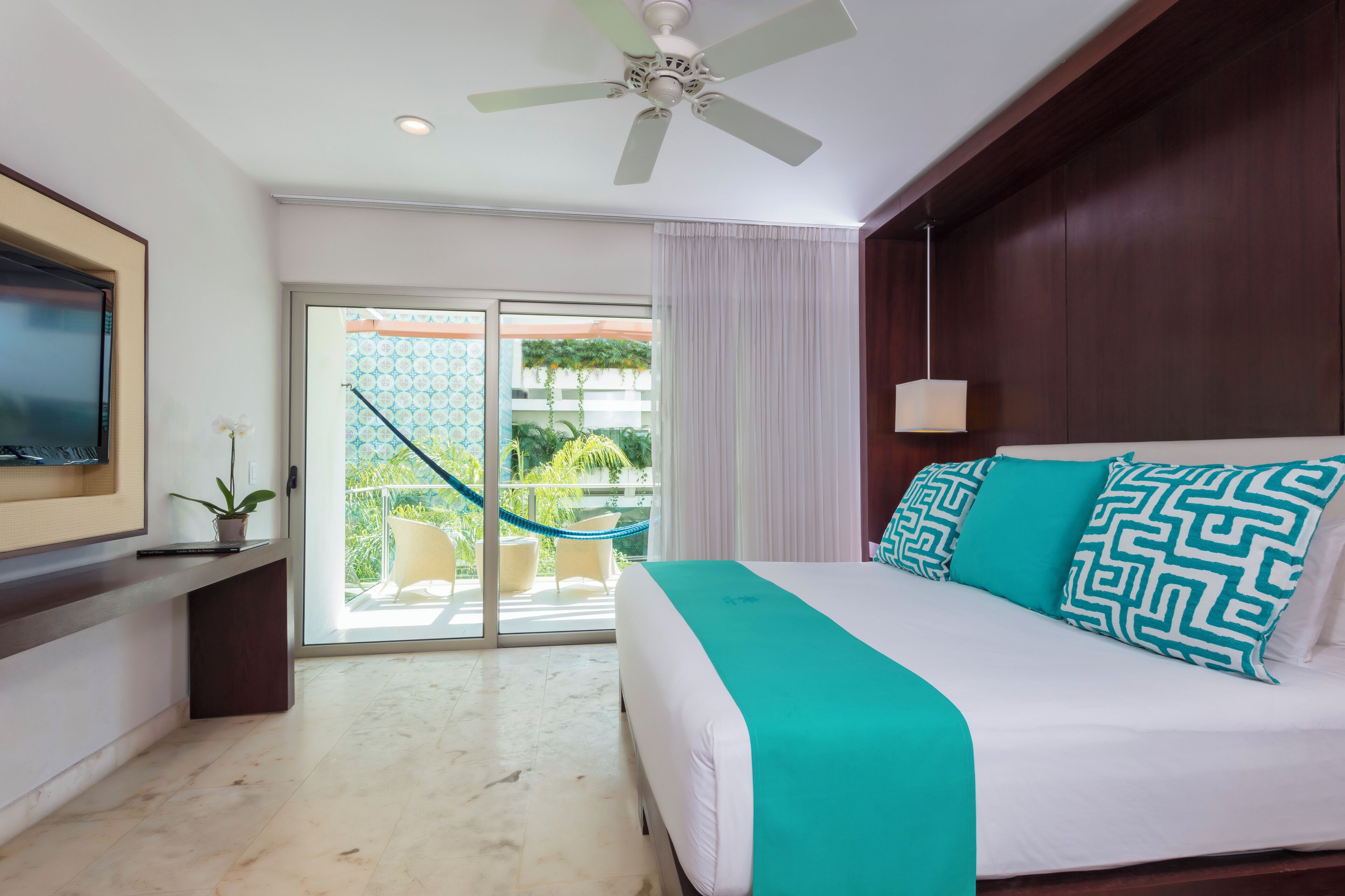 Quarto de hotel com uma cama, um ventilador de teto, uma TV fixada na parede e vista para o exterior através de uma porta deslizante.