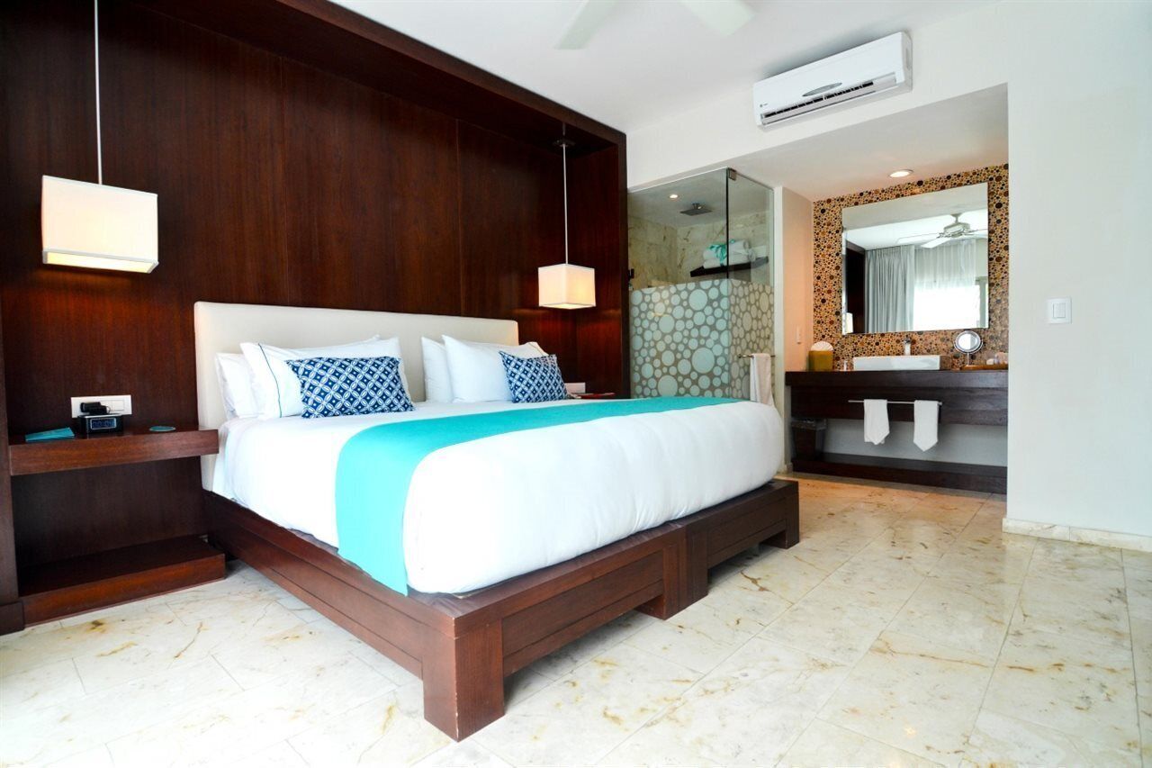 Habitación de hotel moderna con una cama grande, cabecera de madera, banca integrada, tocador con espejo y zona de ducha.