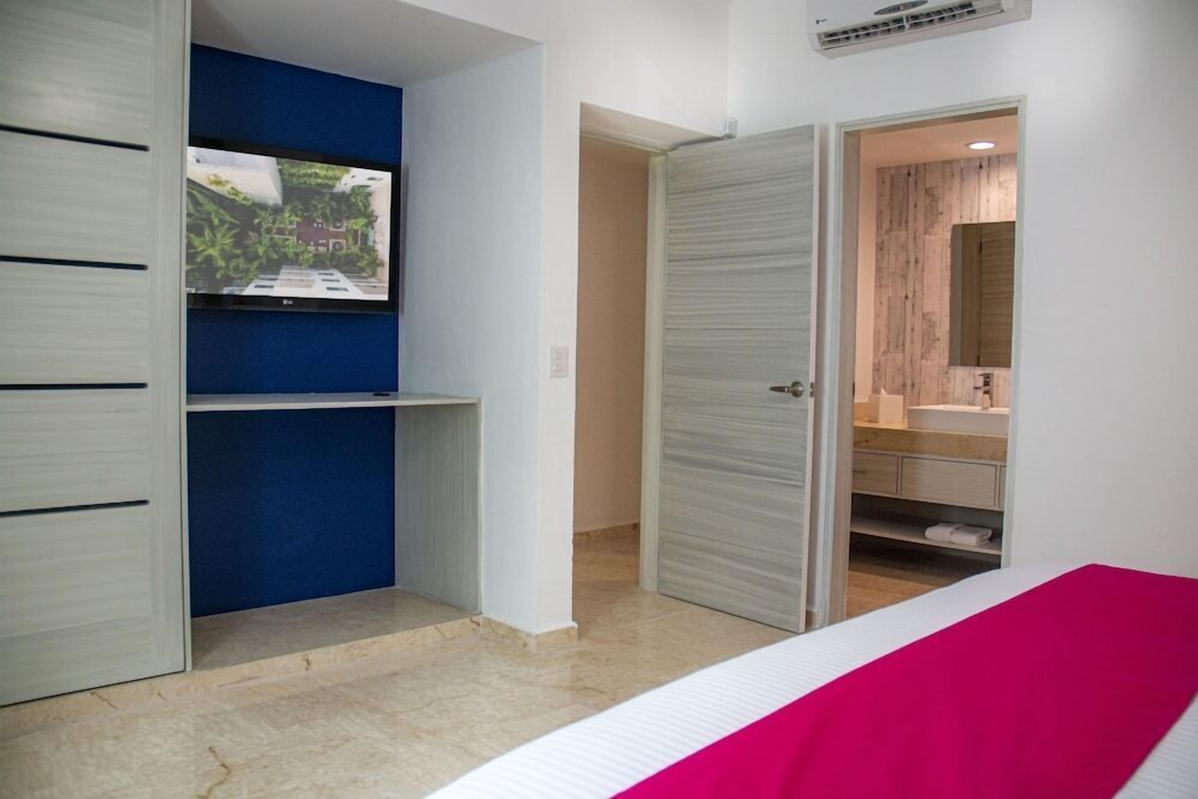 Une chambre à coucher avec un lit, une armoire, un téléviseur et une salle de bain visible à travers une porte ouverte.