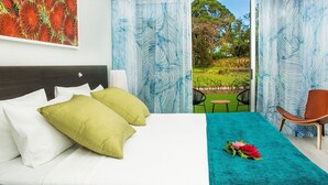 Una camera d'albergo con un letto, due cuscini, una coperta color petrolio, una sedia e vista su un giardino attraverso una finestra.