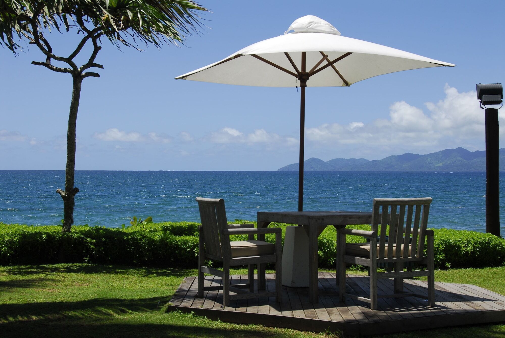 A wooden table and chairs under a white umbrella, overlooking a body of water with mountains in the distance.
