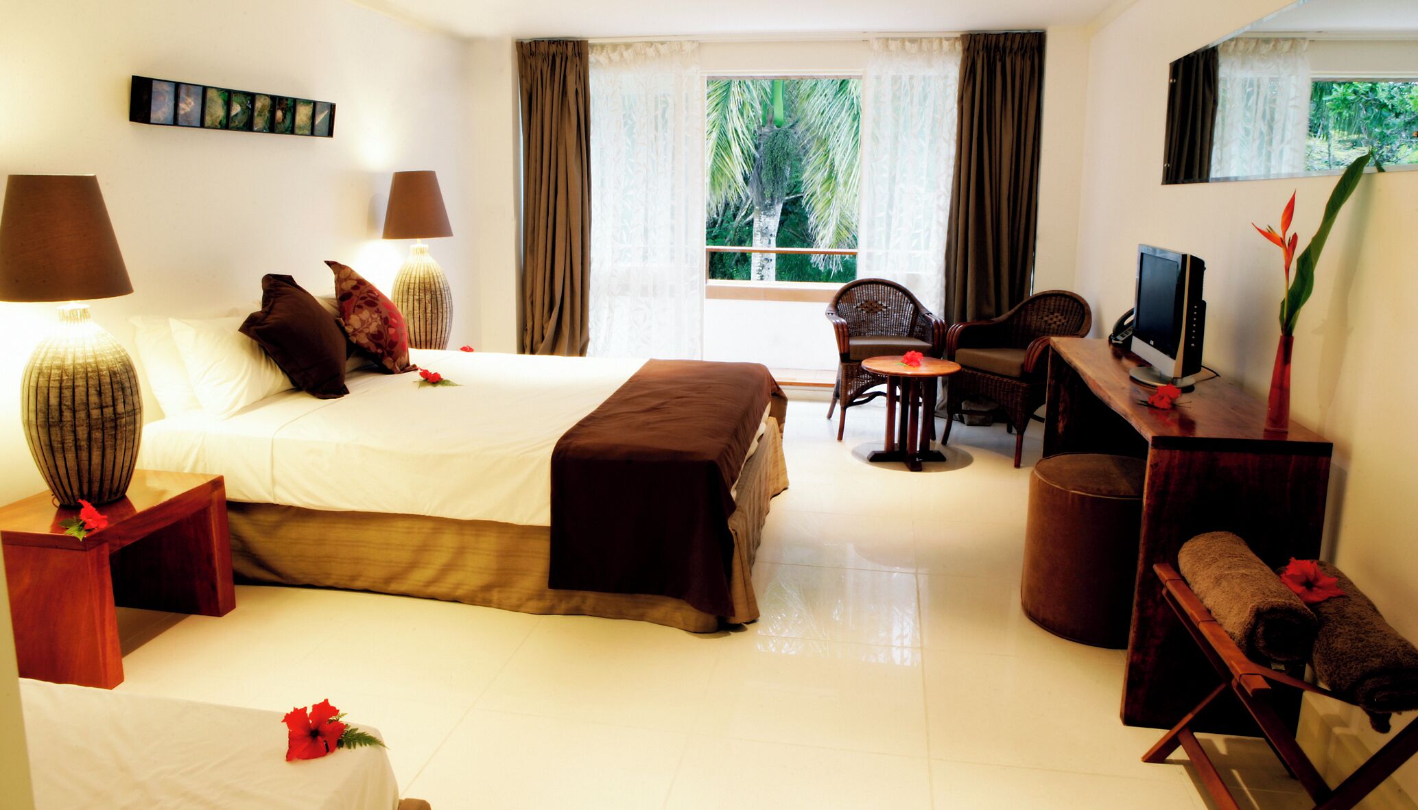 A hotel room with a bed, bedside lamps, a desk with a computer, a chair, a small table with a flower, and a window with curtains.