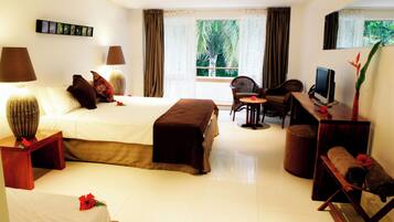 A hotel room with a bed, bedside lamps, a desk with a computer, a chair, a small table with a flower, and a window with curtains.
