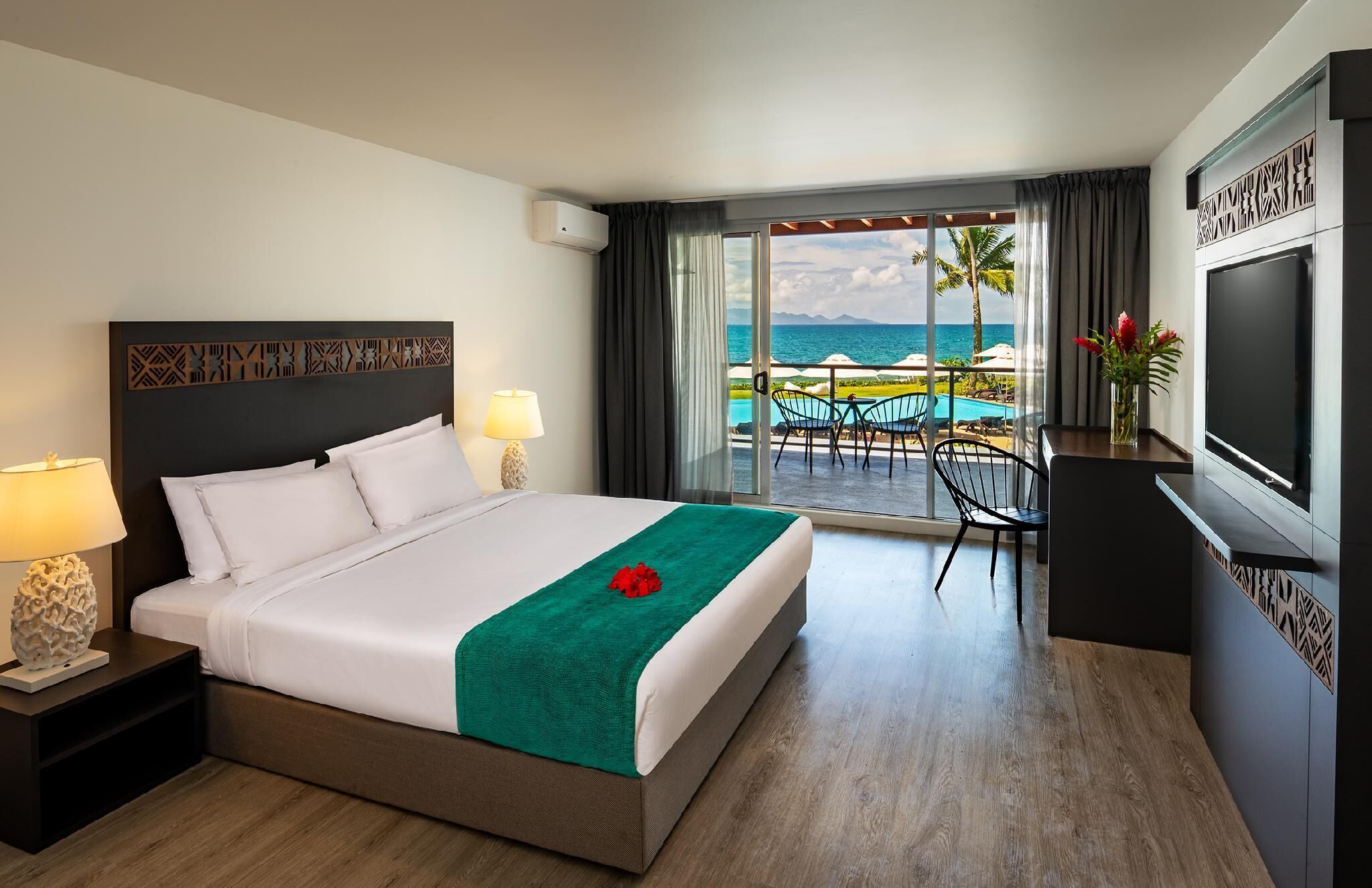 A hotel room with a bed, a TV, a view of the ocean, and a balcony with outdoor furniture.