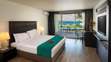 A hotel room with a bed, a TV, a view of the ocean, and a balcony with outdoor furniture.