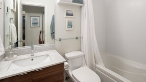 Un bagno con un lavandino bianco, uno specchio e un water. C'è una mensola in vetro sopra il water e una tenda per la doccia.