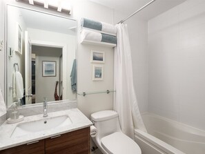 A bathroom with a white sink, mirror, and toilet. There is a glass shelf above the toilet and a shower curtain. - City Oasis - 1br - Furnished 1BR + Parking (Vancouver)