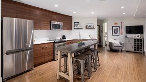 Una cucina moderna con frigorifero in acciaio inossidabile, forno a microonde e piano cottura, mobili in legno, un'isola centrale con sgabelli e un'area pranzo con TV e scaffale per vino.