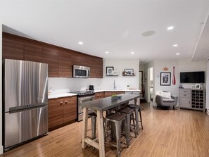 A modern kitchen with a stainless steel refrigerator, microwave, and stove, wooden cabinets, a central island with stools, and a dining area with a TV and wine rack. - City Oasis - 1br - Furnished 1BR + Parking (Vancouver)