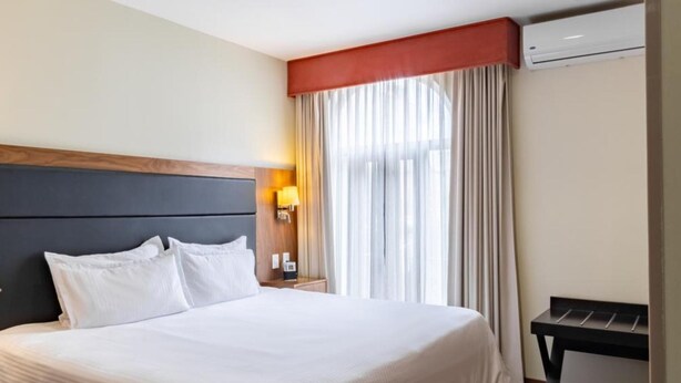 A hotel room with a large bed, a nightstand with a lamp, a window with curtains, and an air conditioning unit. - Senia Reforma (Mexico City)