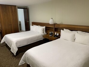 Una habitación de hotel con dos camas, cabecero de madera, una mesita de noche con lámpara y una puerta que da a otra habitación.