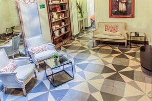 Un hall de hotel con suelo de diseño geométrico, un sofá, sillones, una mesa de centro y una estantería con objetos decorativos.