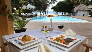 A table set with plates of food, a wine glass, and a vase with flowers, overlooking a pool and beach.